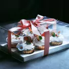 Six chocolate mallow cupcakes topped with white frosting, strawberries, and marshmallows in a clear gift box with a pink ribbon.