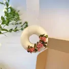 Ring-shaped scented candle with dried pink flowers and green leaves, hanging from a ribbon.