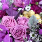 Purple roses, orchids, and carnations arranged with chocolates and greenery.