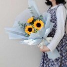 A bouquet of sunflowers and filler flowers wrapped in light blue paper, held by a person in a floral dress.