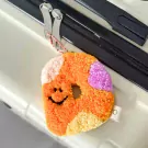 A fluffy donut keychain with a smiley face and colorful patches hangs from a suitcase zipper.
