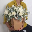 A person in a yellow shirt holds a bouquet of white roses and baby's breath in a brown paper bag vase.