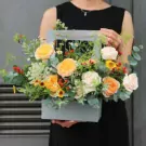 A woman in a black dress holds a pale blue floral arrangement box with various flowers including yellow and pink roses, small yellow sunflowers, and red berries.