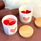 Two white candles in glass jars with red heart wax melts and pink heart labels sit on a pink surface.