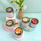 Six scented candles in amber glass jars with labels, featuring various natural ingredients and floral accents.