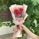Hand holding a bouquet of 3 red roses with delicate fillers, wrapped in light pink and cream paper.