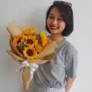 A young woman smiles holding a bouquet of sunflowers wrapped in brown paper and tied with a white ribbon.