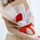 A hand holds a red rose bouquet wrapped in brown paper and white mesh, tied with a red ribbon.