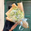 Bouquet of white and light blue flowers with daisies and greenery, wrapped in brown paper and clear plastic with a blue ribbon.