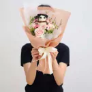 Bouquet with a flower doll on top, pink roses and greenery, wrapped in peach paper, tied with a white ribbon.