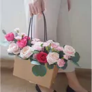 Hand holding a floral arrangement in a brown box with roses and carnations.