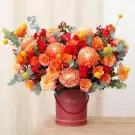 A vibrant bouquet of orange roses, chrysanthemums, and Billy balls in a burgundy box.