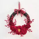 Red rose and berry wreath with ribbon bow hanging on a white wall.