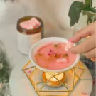 Hand adding pink wax melt cubes to a pink liquid in a white bowl over a candle warmer.