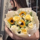 A bouquet of sunflowers, yellow roses, and small white flowers, wrapped in light brown paper.