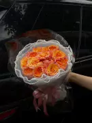 A bouquet of orange and pink roses with white lace trim and pink ribbons, held in front of a black car.