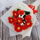 A bouquet of red gerbera daisies and roses with white filler flowers, wrapped in white paper and tied with a red ribbon.