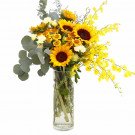 A tall glass vase filled with sunflowers, yellow orchids, and eucalyptus leaves.