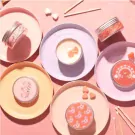 Five pink and peach scented candles in tins arranged on decorative plates, with heart-shaped candies and matches.