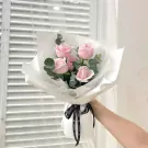 Hand holding bouquet of five pale pink roses, eucalyptus, and baby's breath, wrapped in white paper with Chanel ribbon.