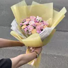 Bouquet of pink and peach roses with purple accents, wrapped in yellow paper, held by two hands.