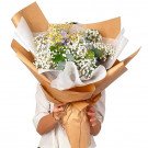 A person holds a bouquet of white baby's breath, yellow daisies, and blue flowers wrapped in brown paper.