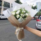 A hand holds a bouquet of white flowers wrapped in burlap and tied with a white ribbon.