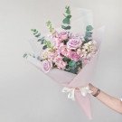 A pastel bouquet of roses, hydrangeas, and snapdragons wrapped in pink paper, held by a person.