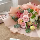 A pastel pink floral bouquet featuring roses, daisies, and other delicate blooms, wrapped in light pink paper.