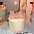 A pale yellow cake topped with a pink bow and a lit candle, adorned with tiny pearls at the base.