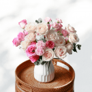 Blushed bouquet of pink and cream roses and carnations in a white vase on a woven tray.
