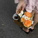 A person holds a brown carrier with a drink and orange flowers, featuring a pink ruffled dress.