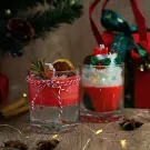 Two Christmas-themed candles in glasses, one with cinnamon stick and dried orange slice, the other topped with whipped cream and festive decorations.