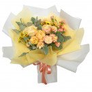 A bouquet of yellow roses, pink carnations, and yellow daisies wrapped in yellow and white paper with a peach ribbon.