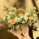Woven basket overflowing with vibrant orange, yellow, and white flowers, held by two hands.