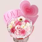 Pink roses and white daisies in a bouquet with a "LOVE" banner and a heart decoration.