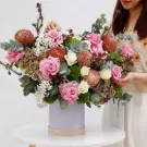 A large floral arrangement in a pastel purple box with pink roses, brown and green chrysanthemums, and other greenery.