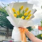 A bouquet of 10 vibrant yellow tulips, wrapped in white paper and tied with an orange ribbon.