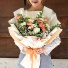 A bouquet of carnations in peach and yellow hues, with baby's breath and greenery, wrapped in cream and peach paper, tied with an orange ribbon.