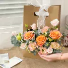 Basket of orange roses, pink roses, and other flowers with eucalyptus in a brown paper bag with a white ribbon.