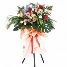 A large flower arrangement on a stand, featuring hydrangeas, roses, gerberas, and tropical leaves.