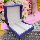 A purple velvet jewelry box with a pearl necklace nestled inside, with a bouquet of pink and white flowers in the background.