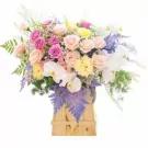 A mixed flower arrangement in a wooden box with pink roses, cream roses, white orchids, and purple accents.