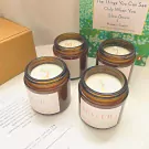 Four amber glass scented candles with white labels on a white surface, with a book in the background.