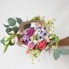 A vibrant bouquet of roses, daisies, snapdragons, and eucalyptus in a brown paper holder.