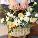 Wicker basket of mixed flowers in peach, yellow, and white, held by hands.