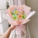 A hand holds a bouquet of colorful hair clip flowers wrapped in pink and white paper, tied with a sheer pink ribbon.