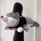 A person holds a large, grey plush husky with a grumpy expression, wearing a black shirt.