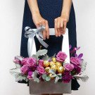Floral arrangement in a silver box with purple roses, Ferrero chocolates, and silver ribbon.