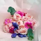 Pink roses, pink gerberas, and blue accents in a bouquet, wrapped in light pink paper.
